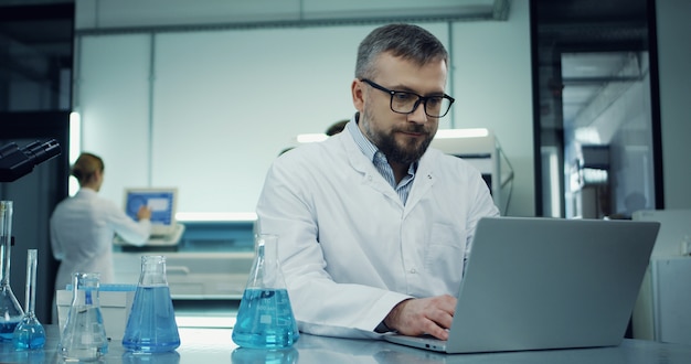 Photo portrait shot of the caucasian man in glasses and white robe working at the laptop computer in the laboratory next with a microscope.