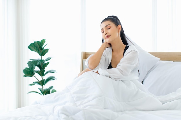 Portrait shot of Millennial Asian sexy female in casual comfort tanktop and shirt sitting under white thick warm blanket posing look at camera smiling after waking up in bedroom in morning at home