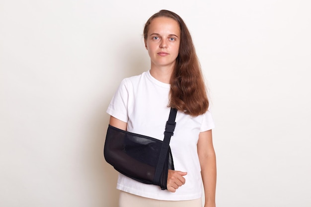 Portrait of sick serious woman wearing casual t shirt injured or broken arm wears splint being on her way home after visit traumatologist posing isolated over white background