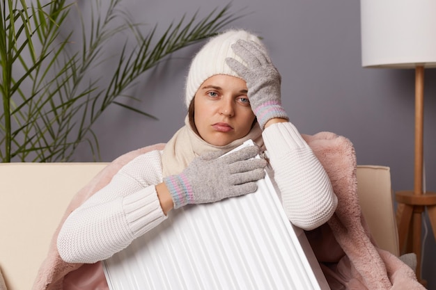 Portrait of sick unhealthy woman in coat and hat sit in cold living room with radiator has no heating in her house suffering flu and headache looking at camera with sad expression