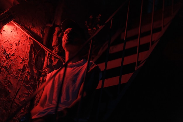 Portrait of sitting on stair adult man with colored red light and dark background.