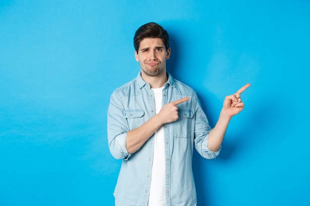 Portrait of skeptical adult guy pointing fingers right and smirking, exress disappointment and doubt, standing against blue wall