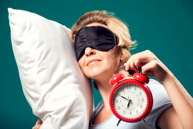 A portrait of sleeping woman with short blond hair with sleep mask on head holding alarm clock and pollow.