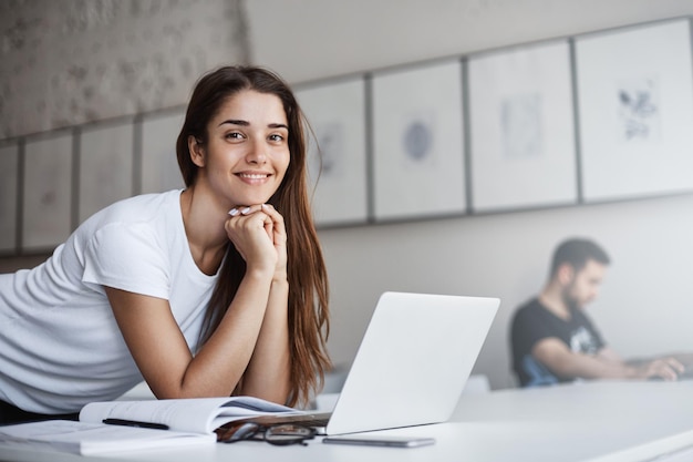 Portrait of sleepy and happy young woman using laptop computer in the morning in busy open space cafe Looking at camera