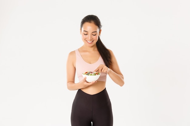   Portrait of slim cute asian fitness girl in sportswear, enjoying eating healthy fresh salad after productive workout 