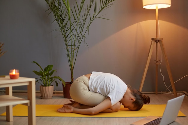 Portrait of slim sporty woman with hair bun wearing white t shirt practicing yoga at home in cozy room posing on floor in child's pose relaxing with online course