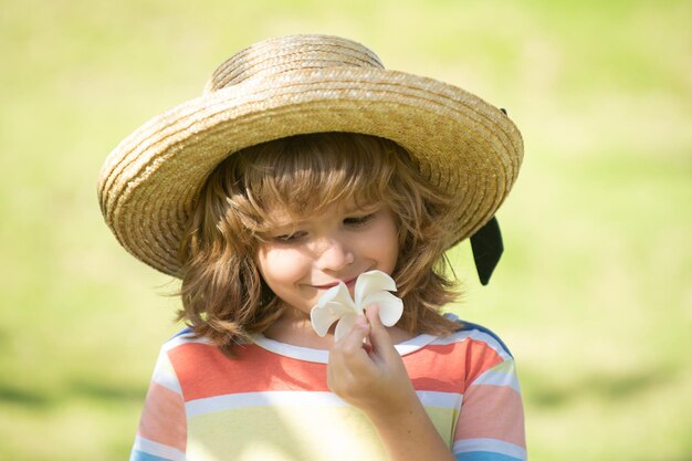 Portrait of a small blond boy in straw hat, closeup. Cute kids face with plumeria flower. Positive emotional child in summer nature park. Summer holiday.