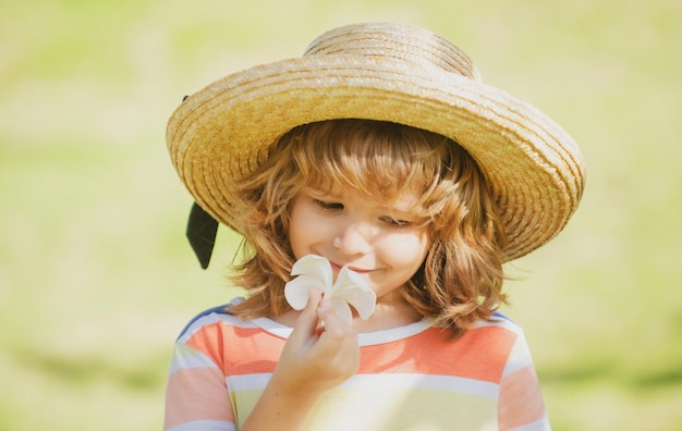 Portrait of a small blond boy in straw hat closeup Cute kids face with plumeria flower Positive emotional child in summer nature park Summer holiday