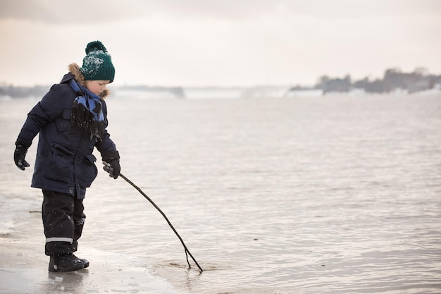 A portrait of a small boy in warm clothes playing with a wooden stick at the river shore