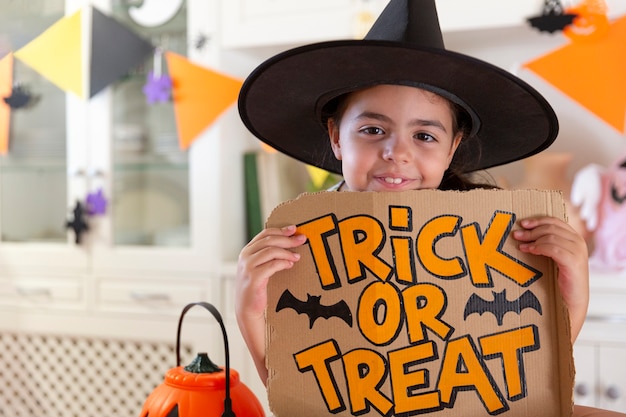 Photo portrait of small caucasian child in witch hat holding a sign with hand drawn trick or treat text.