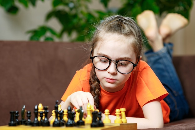 Portrait of a small Caucasian girl with glasses playing chess at home during quarantine due to coronavirus.