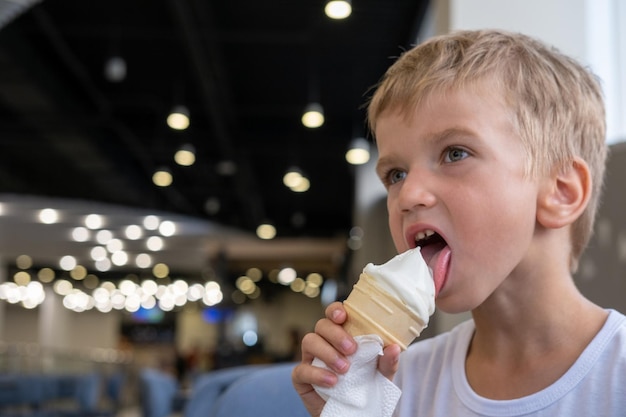 Portrait of a small hungry funny boy eating cold delicious ice cream in a waffle cup sitting in a cafe dark background