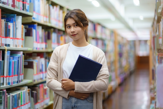 Portrait of Smart Asian woman university student reading book and looking at camera between bookshelves in campus library with copyspace.
