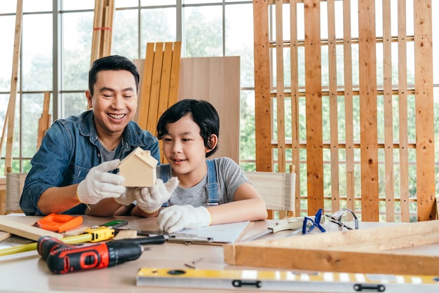 Portrait of smile Asian father and son carpentry sitting building and holding a little wooden home together at home woodwork studio with the instrument of measure and working tools on the table