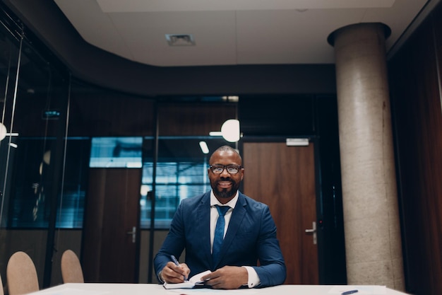 Portrait smiling african american businessman in blue suit sit at table for meeting in office with notebook with pen and laptop