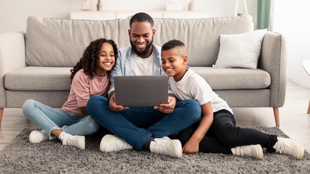 Portrait of smiling African American man and his little children using laptop together, watching movie or having online videocall with mum, browsing internet, sitting on floor carpet in living room