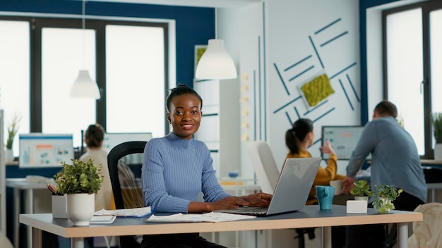 Portrait of smiling african american woman working on profit margin and sale statistics using laptop sitting at desk in busy startup office. Casual business employee happy with job result.