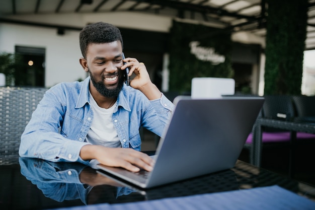 Portrait of smiling african man talking on cell phone while sitting at a cafe with a laptop