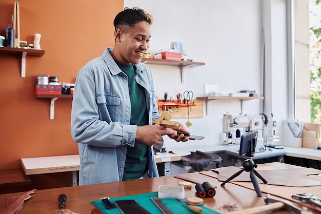 Portrait of smiling artisan recording video tutorial in leatherworking shop and promoting small busi