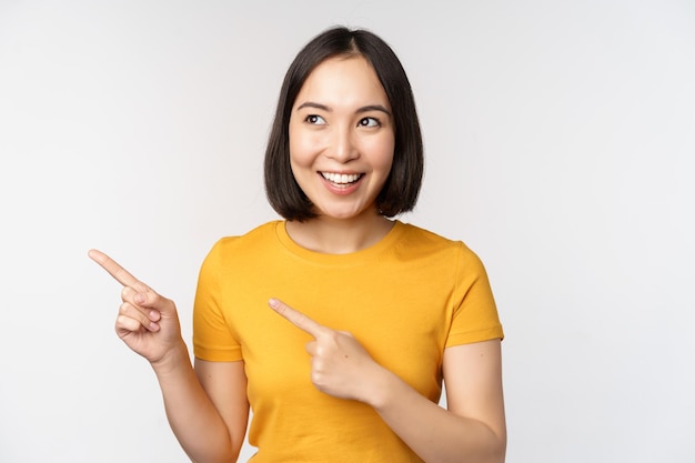 Portrait of smiling asian brunette girl in yellow tshirt pointing fingers left showing copy space promo deal demonstrating banner standing over white background