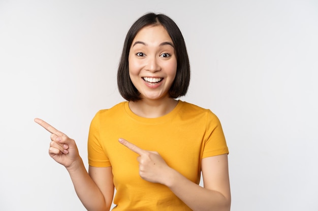 Portrait of smiling asian brunette girl in yellow tshirt pointing fingers left showing copy space promo deal demonstrating banner standing over white background