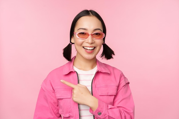 Portrait of smiling asian girl in stylish outfit sunglasses pointing finger left showing advertisement banner standing over pink background