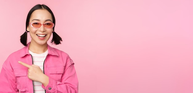 Portrait of smiling asian girl in stylish outfit sunglasses pointing finger left showing advertisement banner standing over pink background