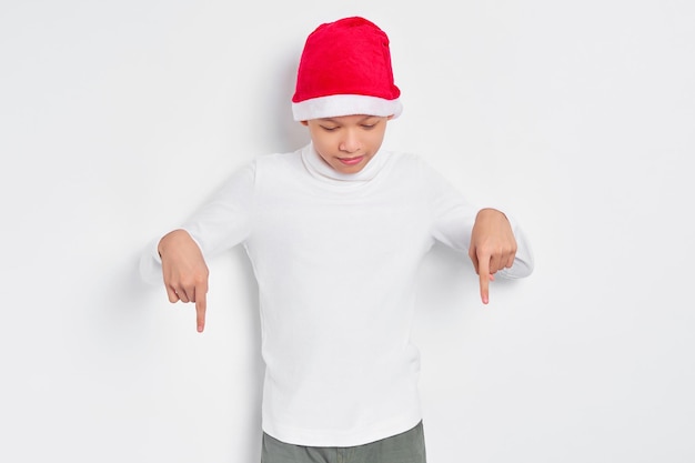 Portrait of smiling Asian man in christmas hat pointing fingers down inviting people to follow isolated on white background