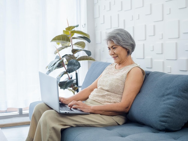 Portrait of smiling Asian senior casual woman sitting on the sofa working with laptop computer in bright white room Elderly female surf internet or video call at home Older people with technology