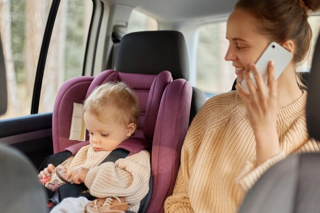 Portrait of smiling attractive mother using phone while sitting in car with her infant daughter on backseat beautiful woman wearing sweater talking on cell phone ans looking at her baby