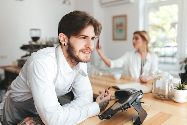 Portrait of smiling barista in apron and white shirt on workplace at cafe. Barista thoughtfully looking aside at the counter while nice girl on background using her mobile phone in coffee shop