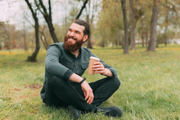 Portrait of smiling bearded man sitting in park on grass and drinking cup of coffee