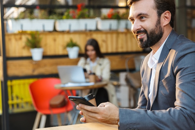 Portrait of smiling bearded man wearing jacket drinking takeaway coffee and using mobile phone while sitting in cafe