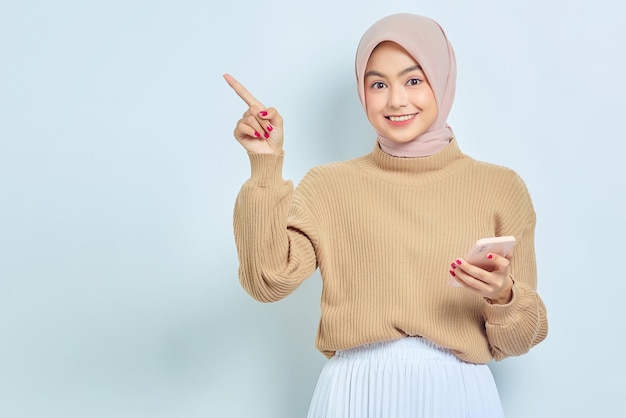 Portrait of smiling beautiful Asian muslim woman in brown sweater and hijab holding mobile phone and pointing finger aside isolated over white background People religious lifestyle concept