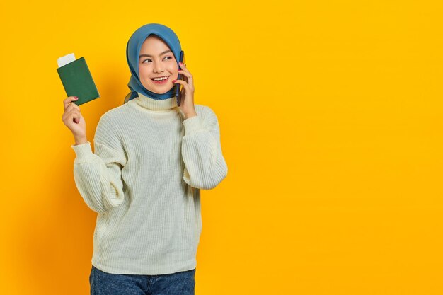 Portrait of smiling beautiful Asian woman in white sweater talking on mobile phone and holding passport looking aside at copy space isolated over yellow background Summer Trip