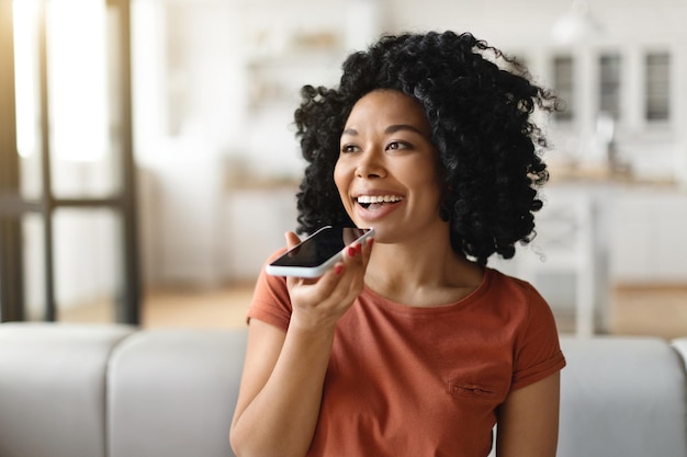 Portrait of smiling black female recording voice message on smartphone at home