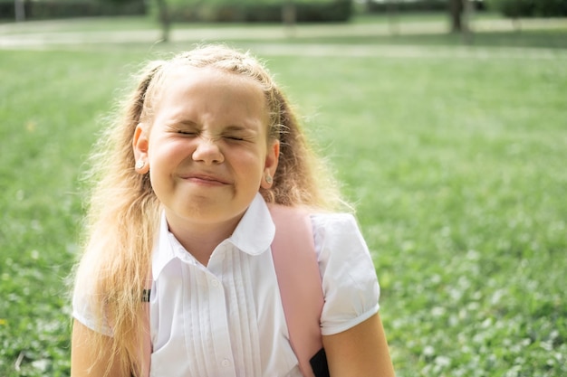 portrait of smiling blonde schoolgirl with close eyes in white shirt with backpack back to school