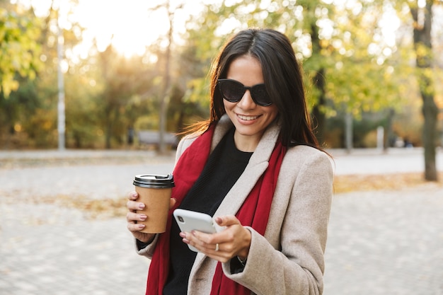 Portrait of smiling brunette woman wearing sunglasses drinking takeaway coffee and typing on cellphone while walking in autumn park