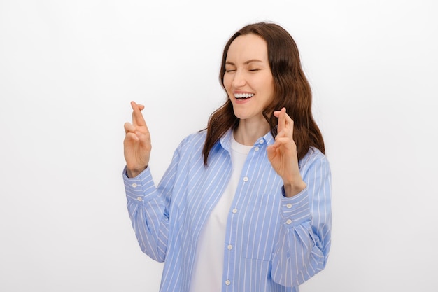 Portrait of smiling brunette woman with closed eyes and with fingers crossed she makes wish in blue shirt on white background for advertising success miracle copy space template