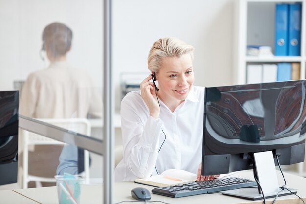 Portrait of smiling businesswoman speaking to microphone while using computer in office interior, customer support concept