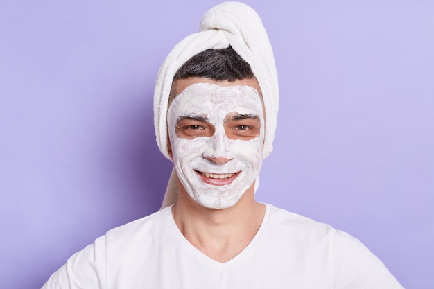 Portrait of smiling Caucasian man with mask for skin standing wrapped in towel isolated over violet background doing cosmetology procedures at home