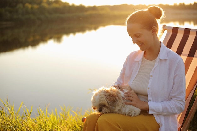 Portrait of smiling Caucasian young adult woman wearing white shirt and her Pekingese dog sitting at sunset near the river petting her favorite animal