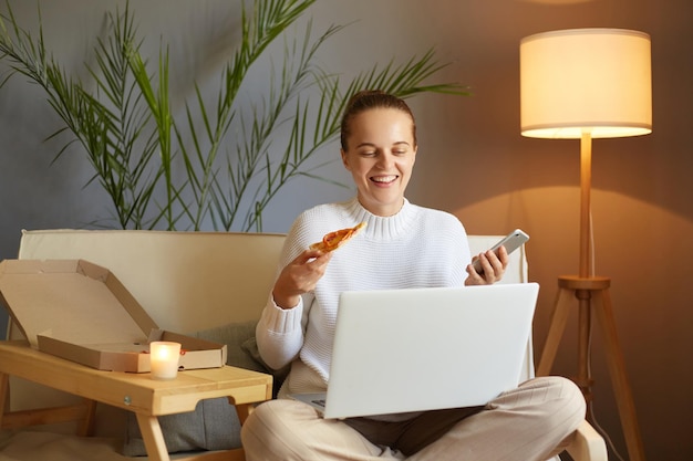 Portrait of smiling Caucasian young adult woman wearing white sweater sitting on sofa and working on laptop holding in hands slice of pizza and mobile phone