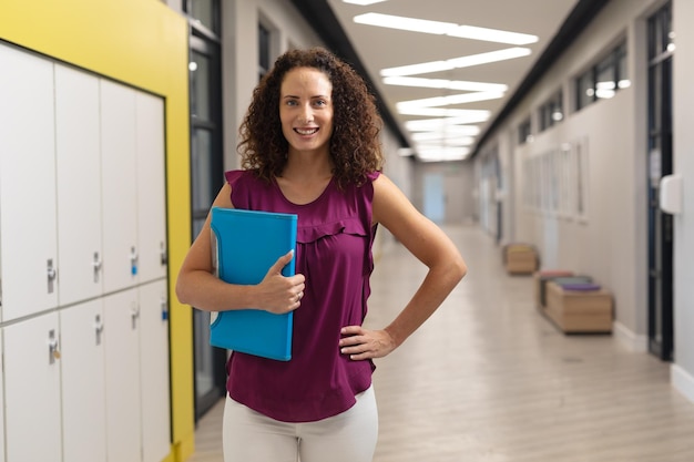 Portrait of smiling caucasian young female teacher with hand on hip standing by lockers in school