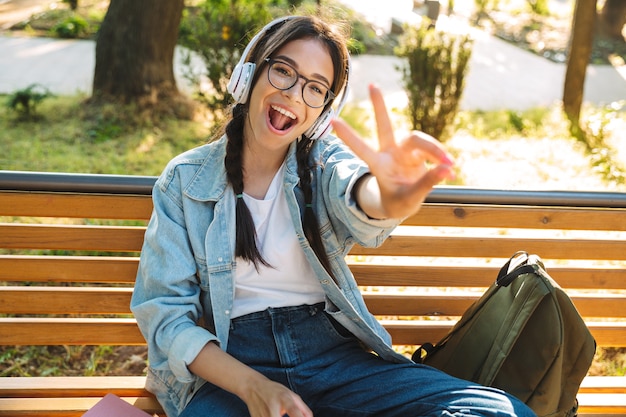 Portrait of a smiling cheerful cute young student girl wearing eyeglasses sitting on bench outdoors in nature park listening music with headphones showing peace gesture.