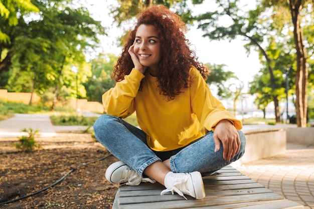 Portrait of a smiling cheerful happy young beautiful curly student outdoors in nature park sitting on a bench.