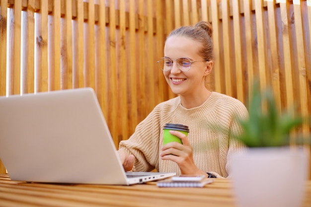 Portrait of smiling delighted satisfied woman freelancer with bun hairstyle wearing beige sweater working on laptop and drinking coffee looking at display with happy expression