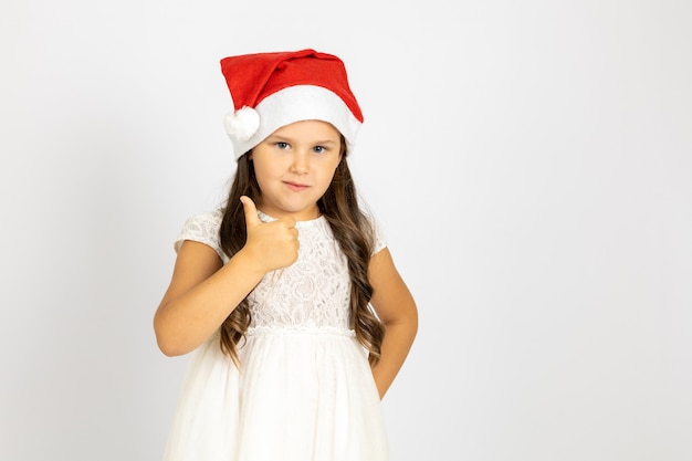 Portrait of smiling enthusiastic child in red santa claus hat gives thumbs up isolated on white back...