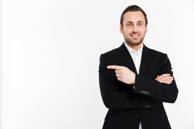 Portrait of smiling entrepreneur having stubble wearing black suit and pointing index finger aside, isolated over whitecopy space