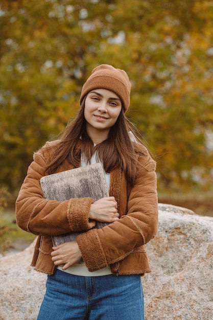 Portrait of smiling female higher education student with laptop standing in park in fall